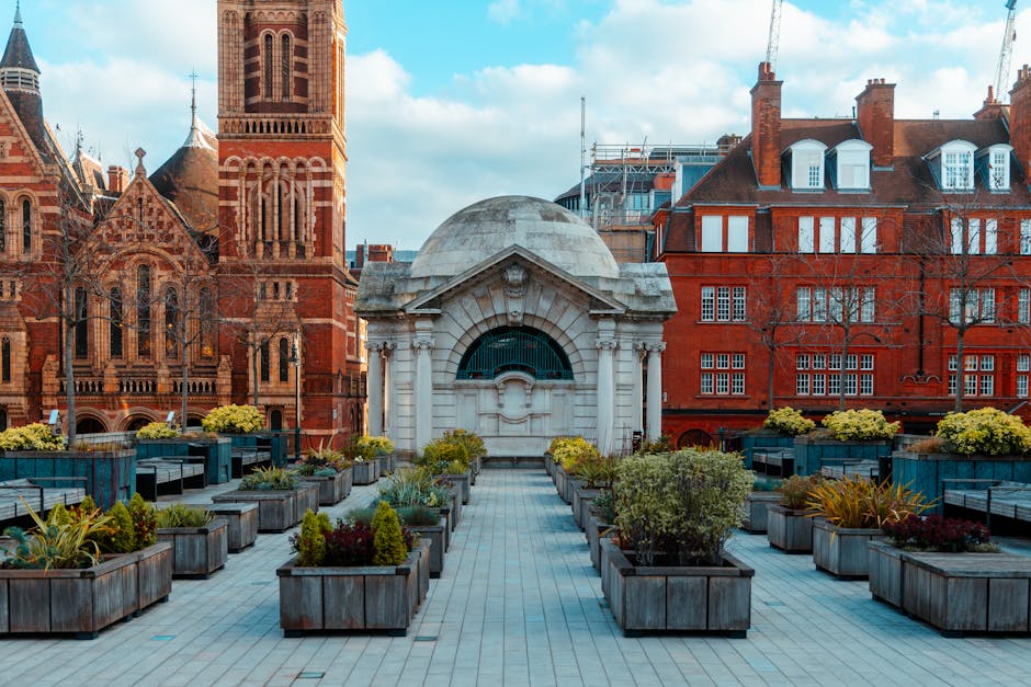 A spacious urban garden terrace situated on a rooftop, featuring a symmetrical layout with paved pathways leading to a central decorative pavilion with a curved, domed roof and intricate stone detailing. Surrounding the pathways are numerous rectangular wooden planters filled with a variety of lush, well-maintained plants, including leafy shrubs, flowering annuals, and small conifers, showcasing vibrant green and subtle red and yellow accents. The garden area is bordered by seating benches and planters, creating a relaxing outdoor space amidst the cityscape. In the background, historic red-brick buildings with ornate architectural features, such as arched windows and decorative stonework, add to the scenic urban environment under a partly cloudy sky. The scene captures the atmosphere of a well-kept city garden, maintained by Gardeners Shepherd's Bush, emphasizing landscaping work, lawn care, and hedge trimming in an aesthetically pleasing setting.