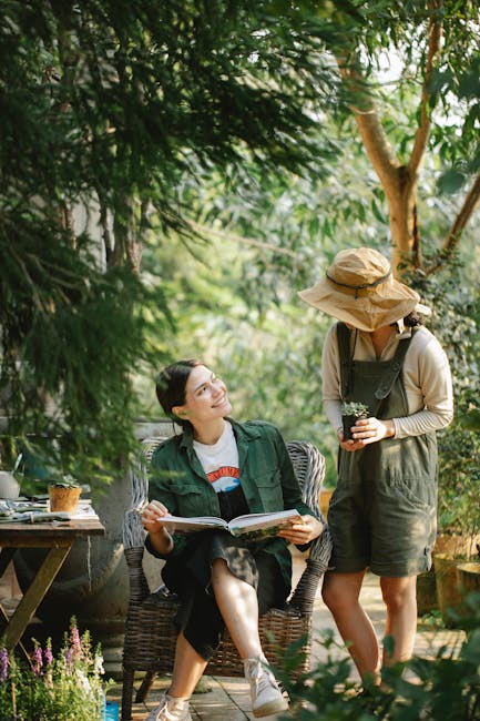 In a lush garden setting with abundant green foliage and tall trees, two women are engaged in gardening activities. One woman with dark hair, wearing a green jacket and casual clothes, is seated comfortably on a wicker chair, holding an open book or magazine in her hands, and looking up smiling at the other woman. The standing woman, dressed in a wide-brimmed straw hat, beige long-sleeve top, and green overalls, is holding a small potted plant, possibly for planting or pruning. The scene features a well-maintained lawn with trimmed grass, garden beds filled with flowering plants and herbs, and a pathway partially visible. To the left, there is a rustic wooden table with gardening tools and a small potted plant, suggesting ongoing gardening or landscaping work. The overall atmosphere is bright and peaceful, capturing a moment of outdoor garden maintenance and planting. The image naturally highlights the gardening activities and the lush environment, aligned with services offered by Gardeners Shepherd's Bush.
