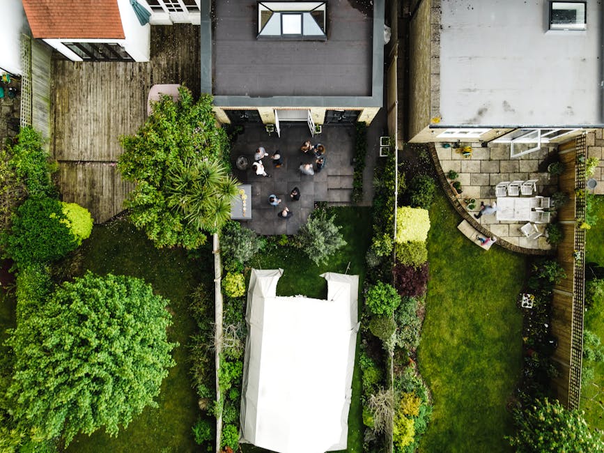 An aerial view of a residential garden showing various landscaping features and outdoor areas. The garden includes a lush, green lawn with well-maintained grass bordered by a wooden fence. To the left, a dense grouping of trees and shrubs provides privacy and natural beauty, with textures ranging from broad leafy canopies to smaller, delicate branches. In the center, a paved patio area hosts a group of seven people standing and conversing, with some sitting on nearby benches. Adjacent to the deck, a garden bed with neatly trimmed bushes and flowering plants outlines the space. On the right, a circular stone patio provides a space for outdoor dining, with a white table and several chairs, surrounded by potted plants and small decorative elements. The background features a modern building with large windows and a flat roof, contrasting with the landscaped garden below. The overall scene reflects careful lawn care, hedge trimming, and garden maintenance, exemplifying the quality of landscaping work that Gardeners Shepherd's Bush offers to maintain vibrant outdoor spaces.